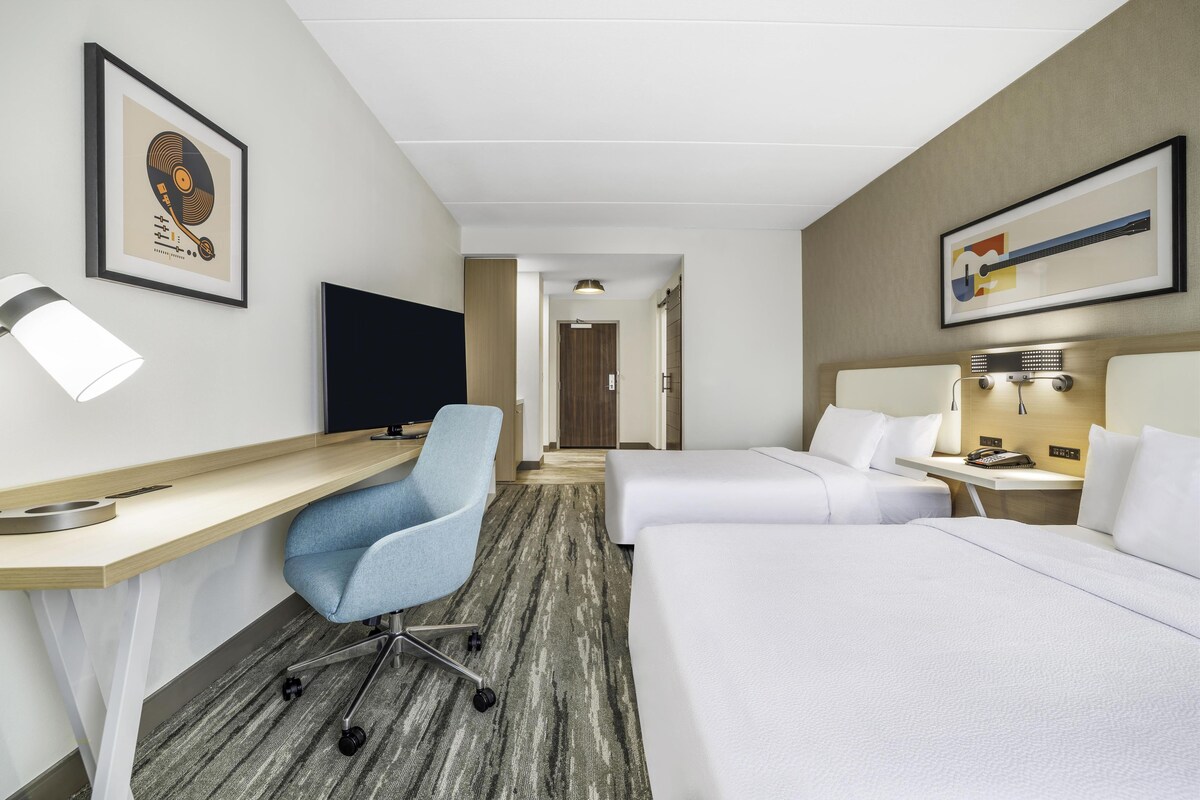 Una habitación de hotel con escritorio, dos camas, televisión y obras de arte en las paredes.