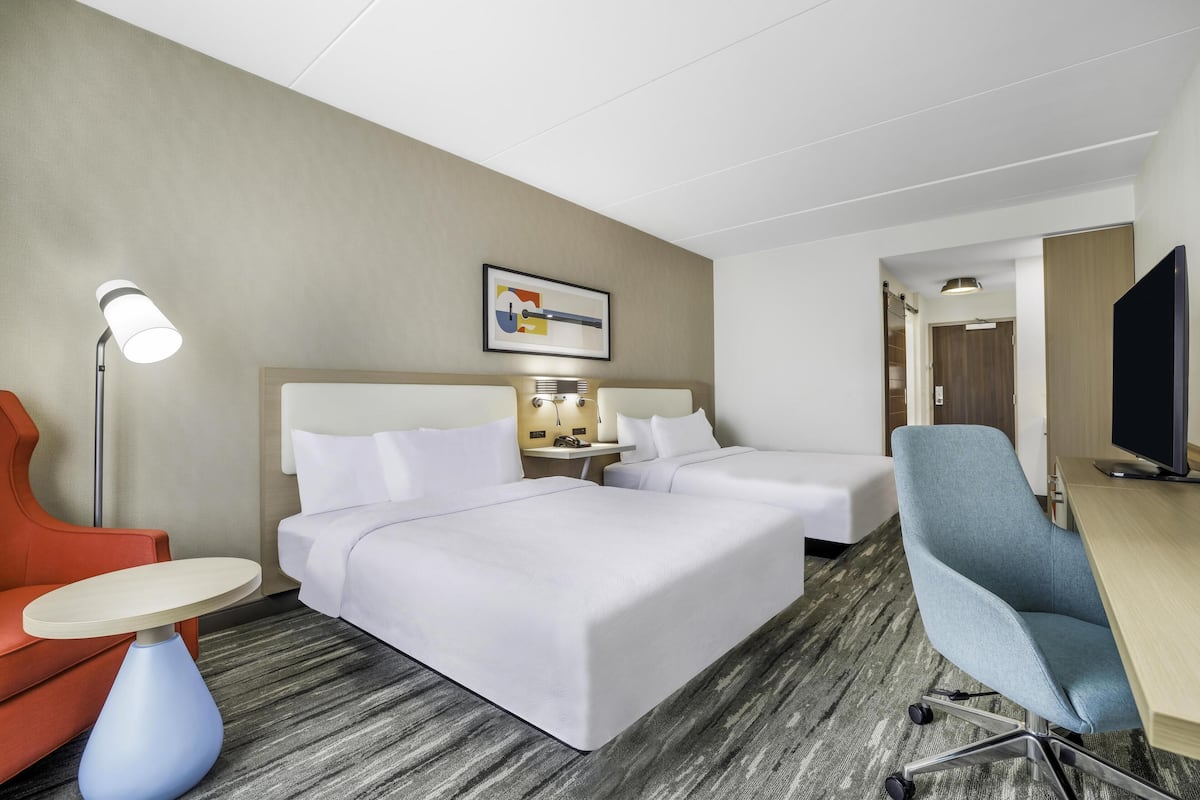 Quarto de hotel com duas camas, uma escrivaninha, uma cadeira, um abajur e uma televisão.