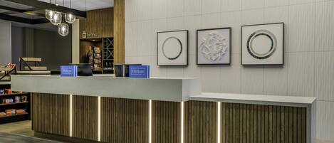 A modern hotel reception area with a wooden reception desk, illuminated by under-cabinet lighting. There are framed artworks on the wall and a 'Hilton' sign above the desk.