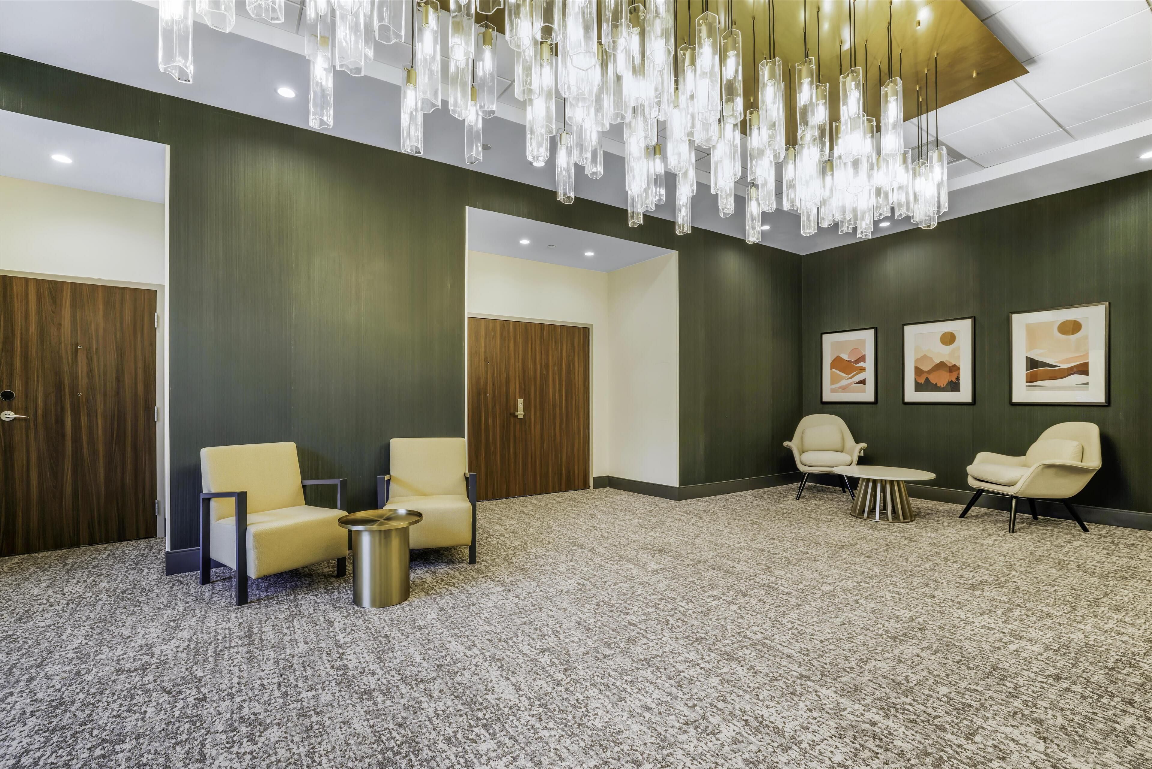 A lobby with a carpeted floor, two yellow armchairs, a small round table, and a chandelier.
