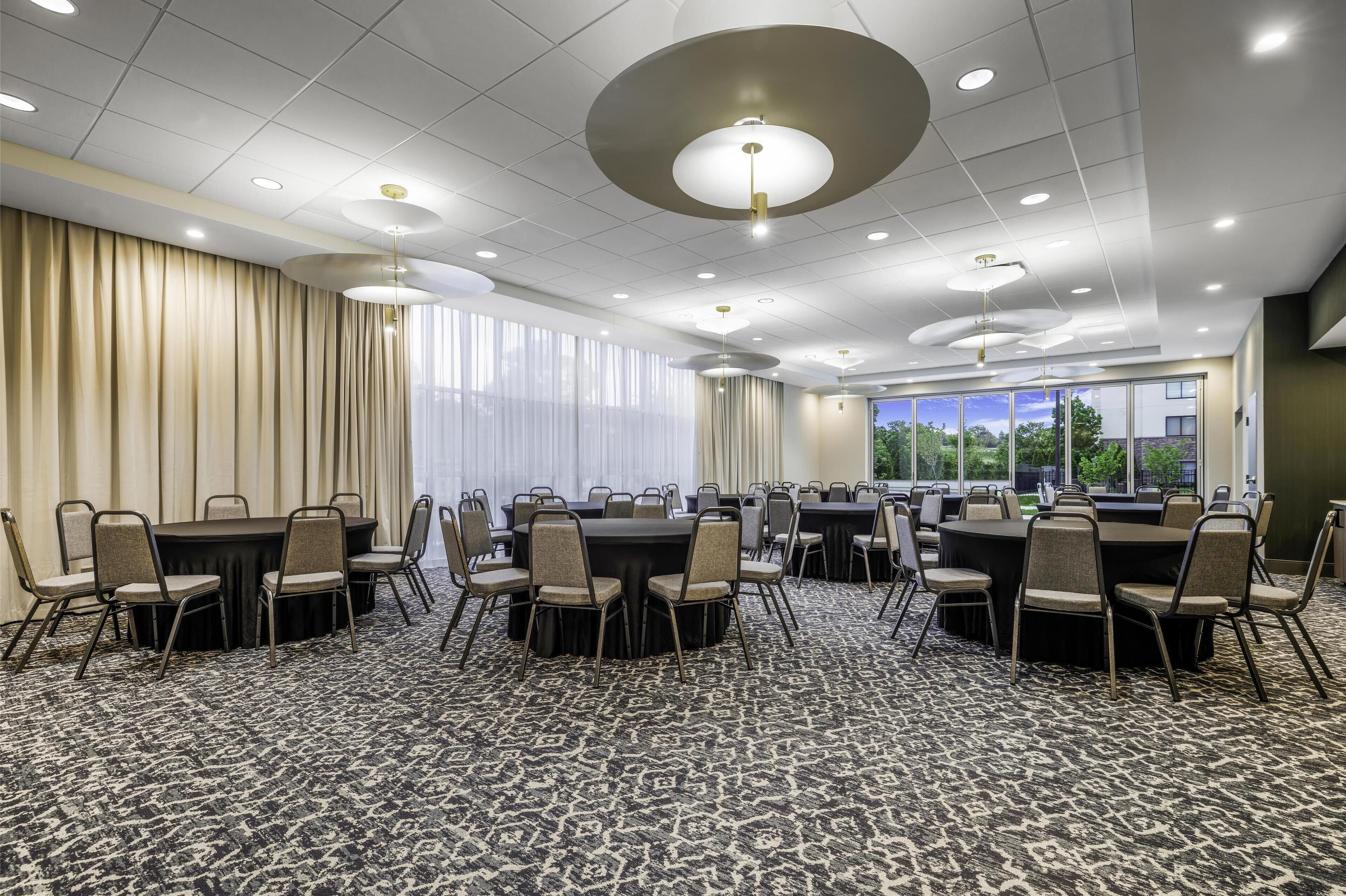 A conference room with multiple tables and chairs, large windows with curtains, and a patterned carpet.
