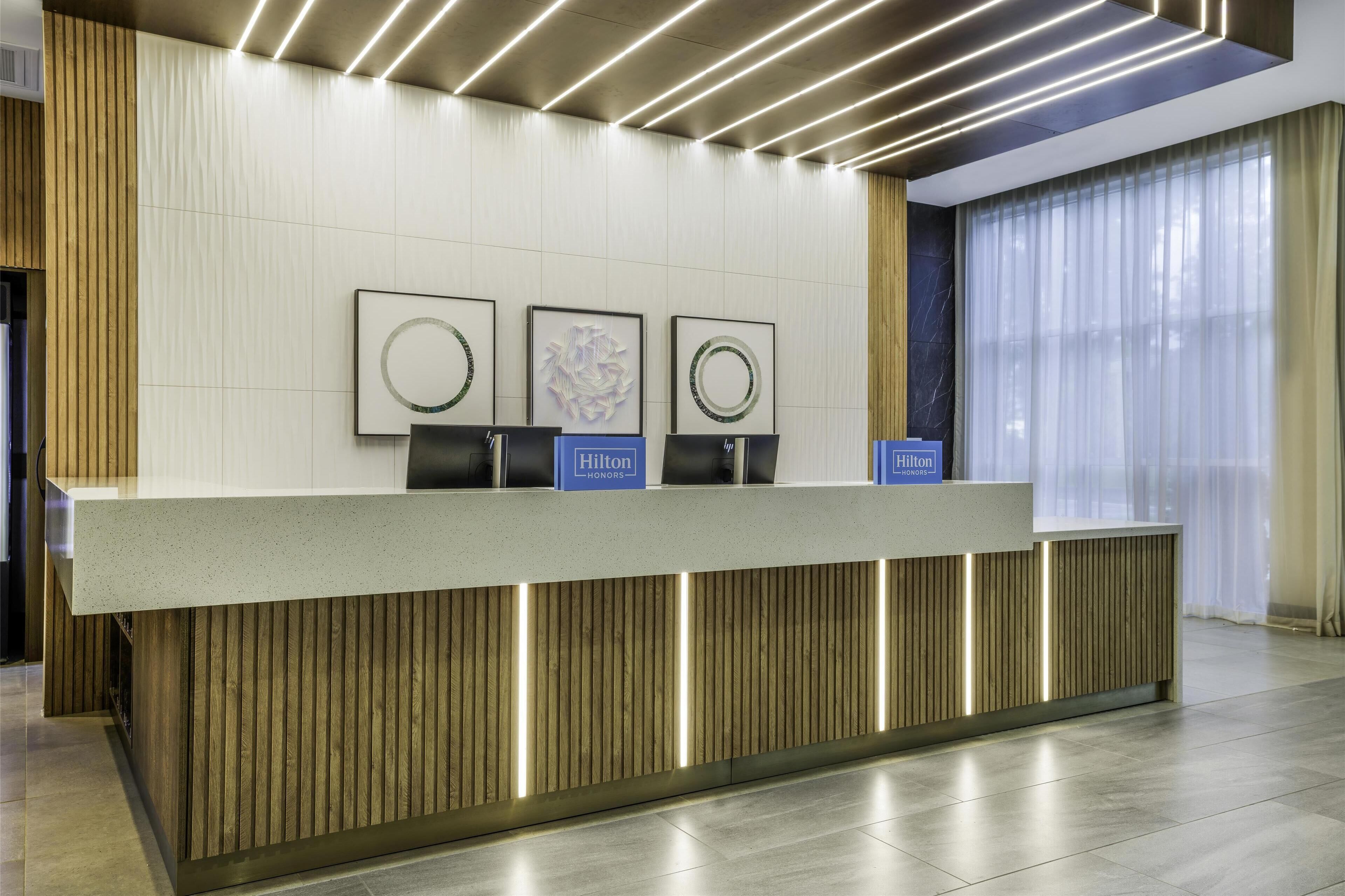 Un moderno mostrador de recepción de hotel con un diseño limpio y minimalista, que incluye un panel de madera decorativo e iluminación en el techo.
