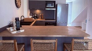 Uma cozinha moderna com armários escuros, micro-ondas e geladeira de aço inoxidável. Há uma ilha de madeira com banquetas, uma bandeja de copos e uma chaleira.