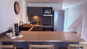A modern kitchen with dark cabinetry, a microwave, and a stainless steel refrigerator. There is a wooden island with bar stools and a tray of cups and a kettle.