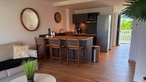 Uma cozinha moderna com uma ilha central, uma área de refeições com cadeiras de vime, uma mesa de centro redonda e um sofá com um almofado decorativo.