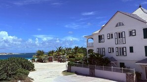 Um edifício branco de dois andares com varanda, vista clara do oceano e um caminho de paralelepípedos até a entrada.