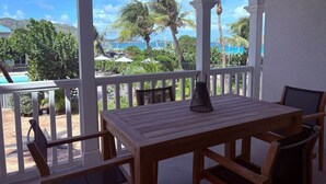 Uma área de estar coberta ao ar livre com mesa e cadeiras de madeira, com vista para uma piscina e palmeiras.