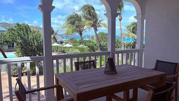 A covered outdoor seating area with a wooden table and chairs, overlooking a pool and palm trees.