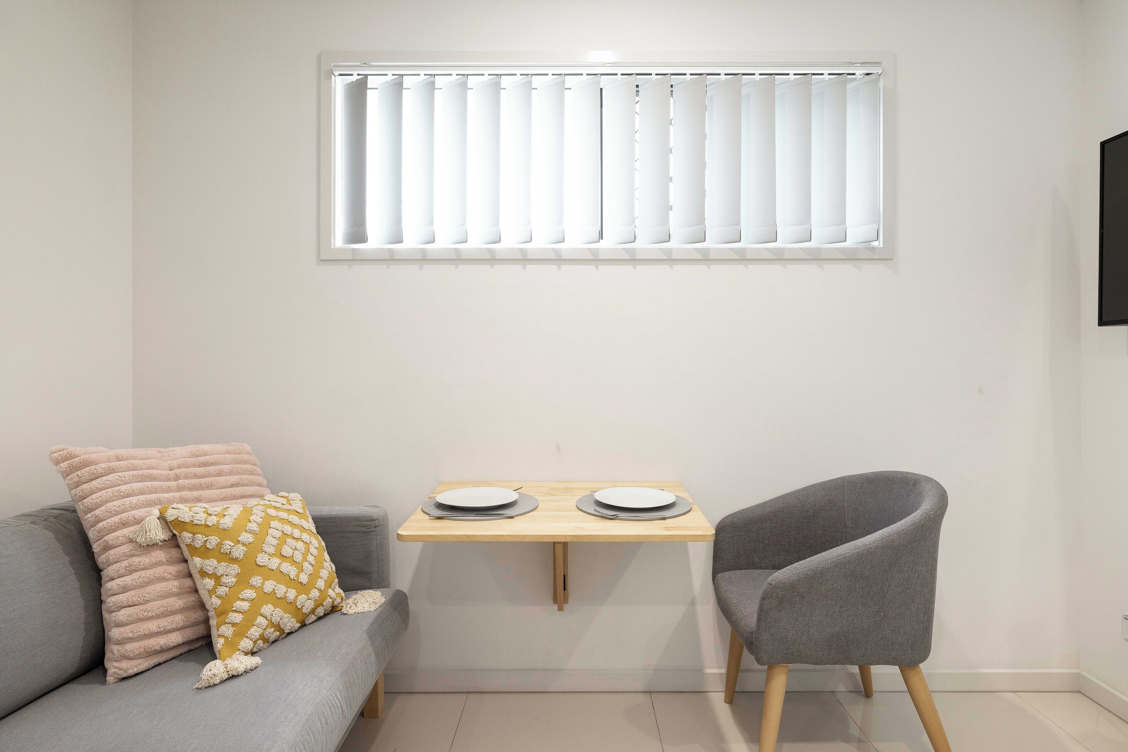 A room with a grey sofa, a small wooden table with two plates, a grey armchair, and a window with blinds.