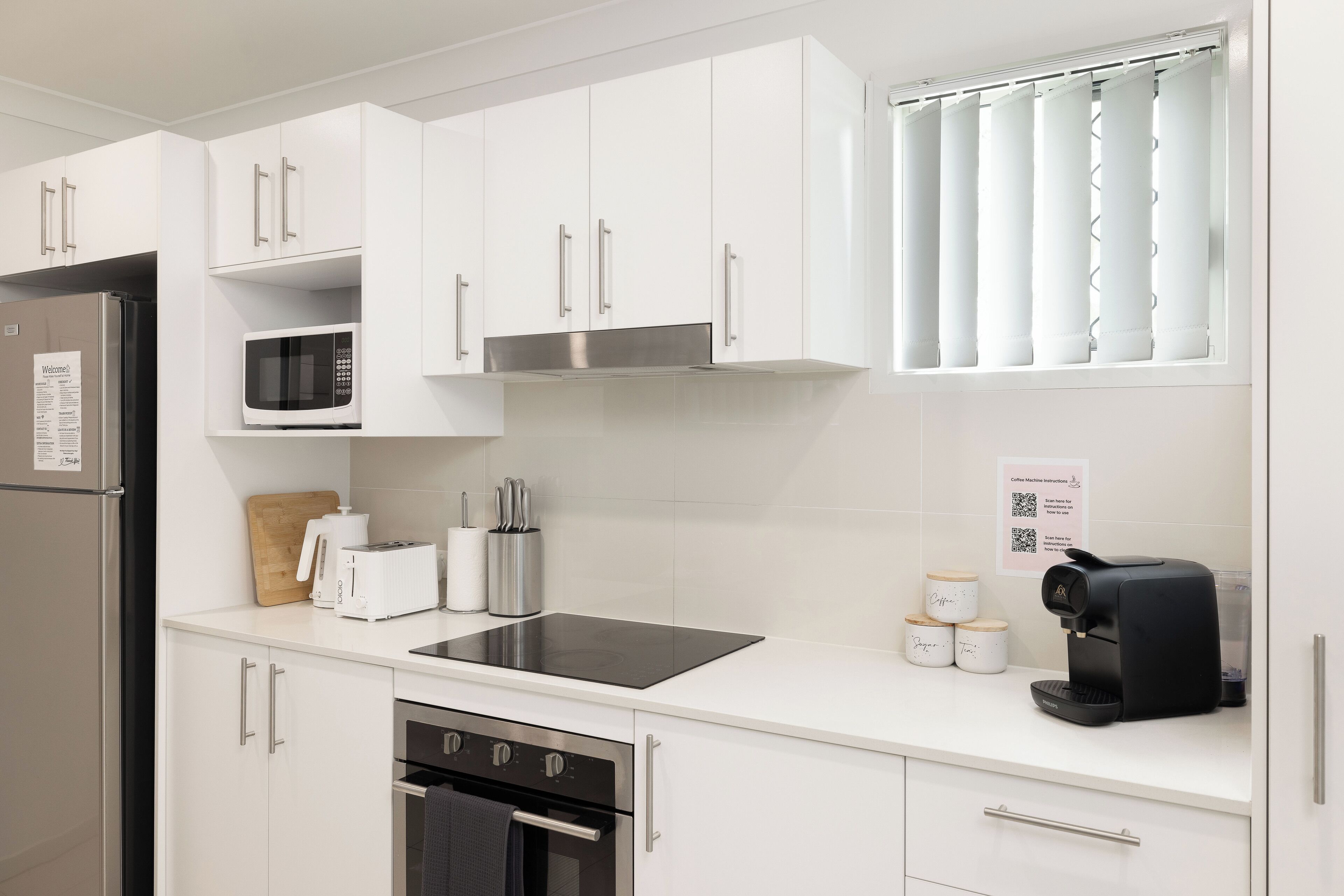 Een moderne keuken met witte kastjes, een roestvrijstalen koelkast, een magnetron, een broodrooster, een koffieapparaat en een inbouwoven.