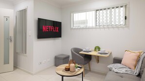 A modern living room with a flat-screen TV displaying "NETFLIX," a gray sofa, a small round table with a vase and remote, and a window with blinds. - Smart Suites Self Check-in Apartments - 39A Freney (Rocklea)