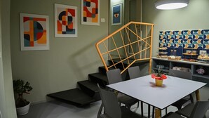 A modern dining area with a white table, grey chairs, a staircase with an orange railing, and abstract art on the walls.