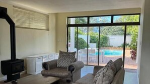A living room with a sofa, armchair, and a fireplace. There is a large window with a view of a swimming pool and greenery outside. - Heaven  (Cape Town)
