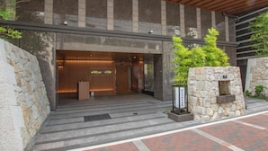 A modern building entrance with a stone facade, a wooden reception desk, and a small stone structure with a water feature. - Sora Niwa Terrace Kyoto (Kyoto)