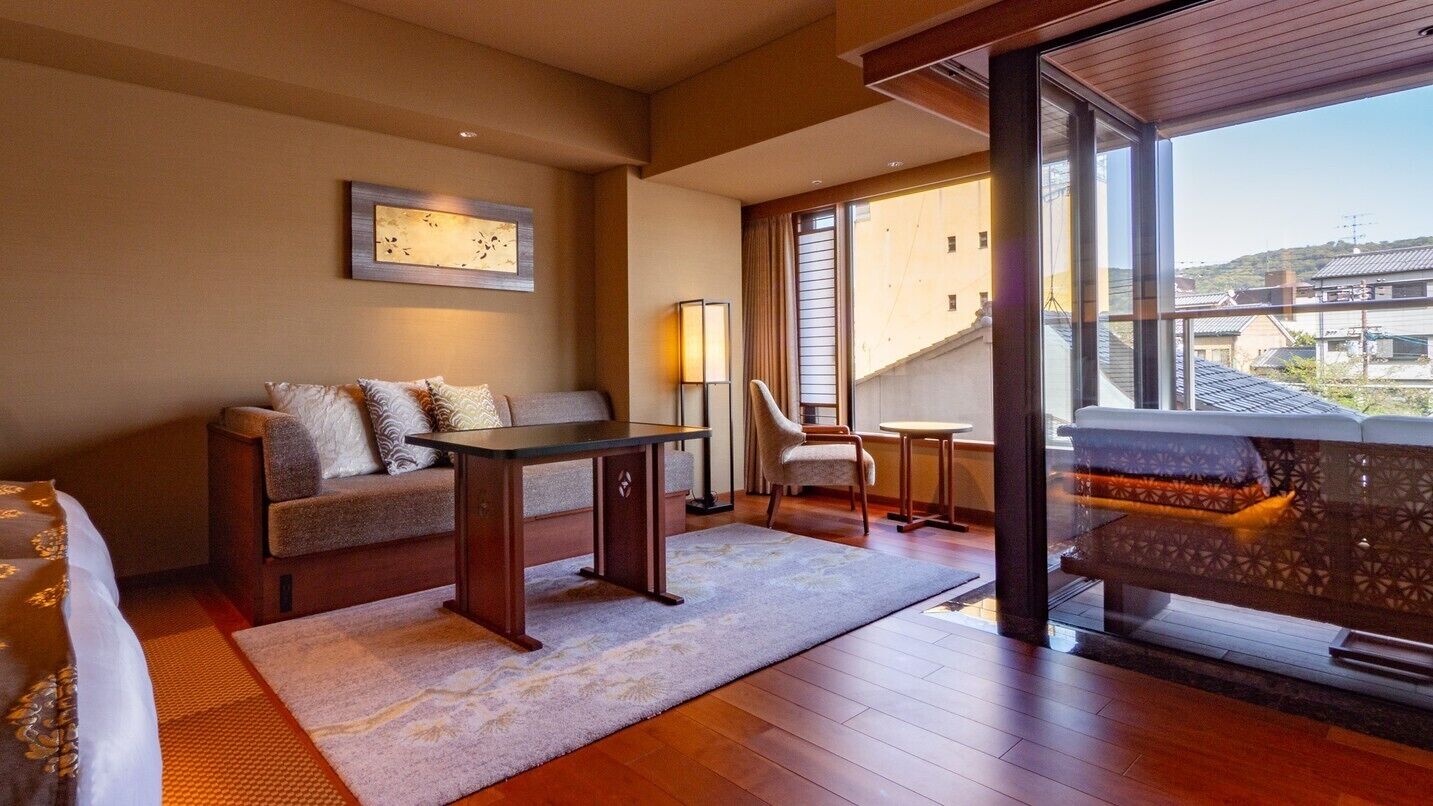 A hotel room with a sofa, a small table, a chair, a lamp, and a view of the outside.