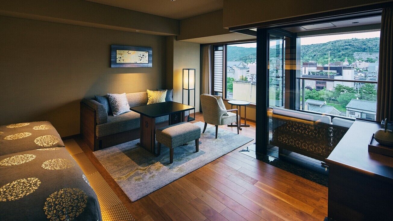 A modern hotel room with a sofa, a small table, a chair, a view of the city through large windows, and a balcony with a railing.