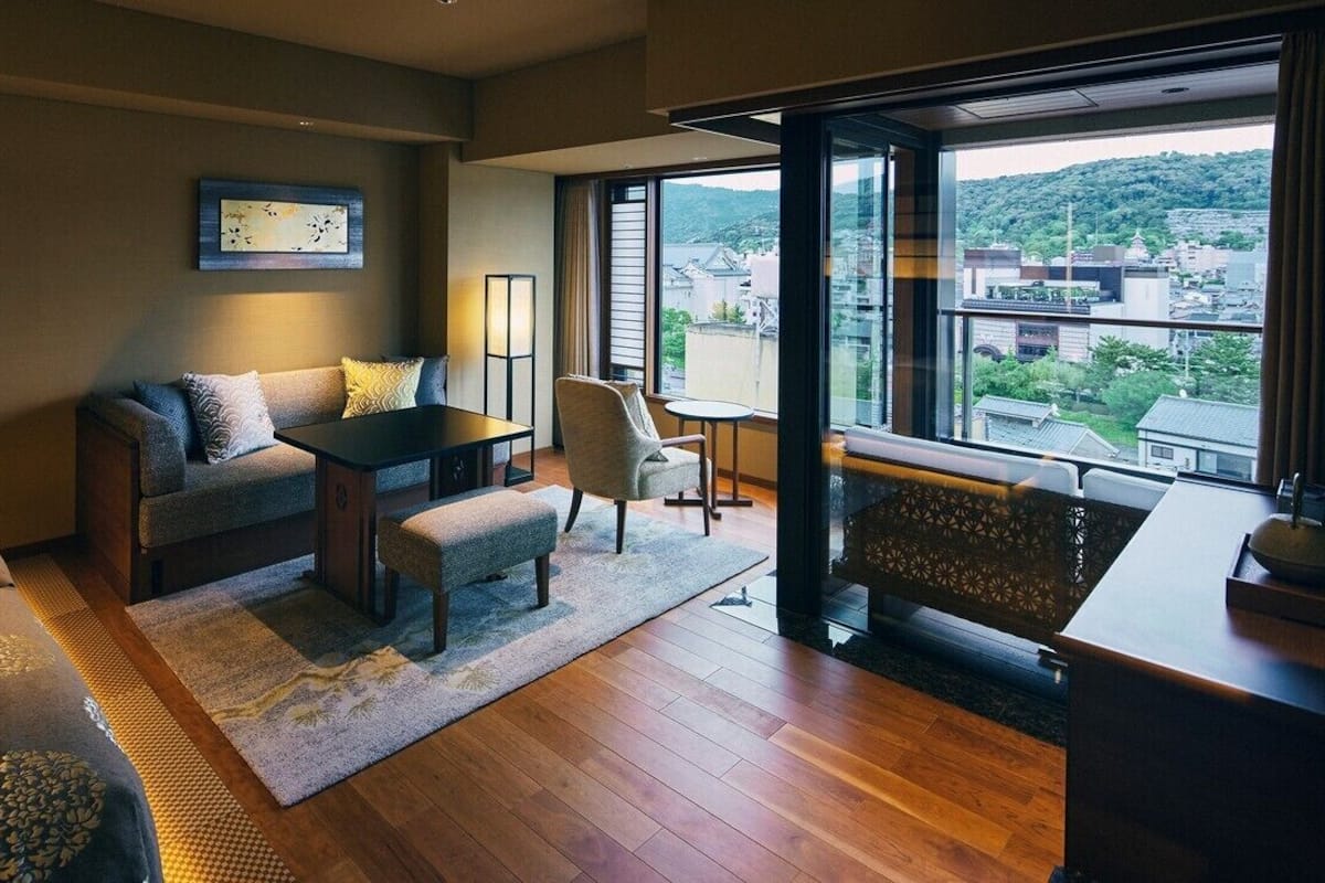 Ein modernes Hotelzimmer mit einer Sitzecke, einem kleinen Tisch, einem Sessel, Blick auf die Stadt durch große Fenster und einem Balkon mit Geländer.