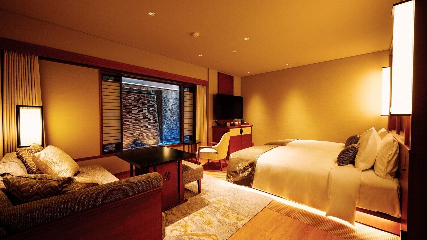 A hotel room with a bed, a sofa, a small table, a TV, and a window with blinds.