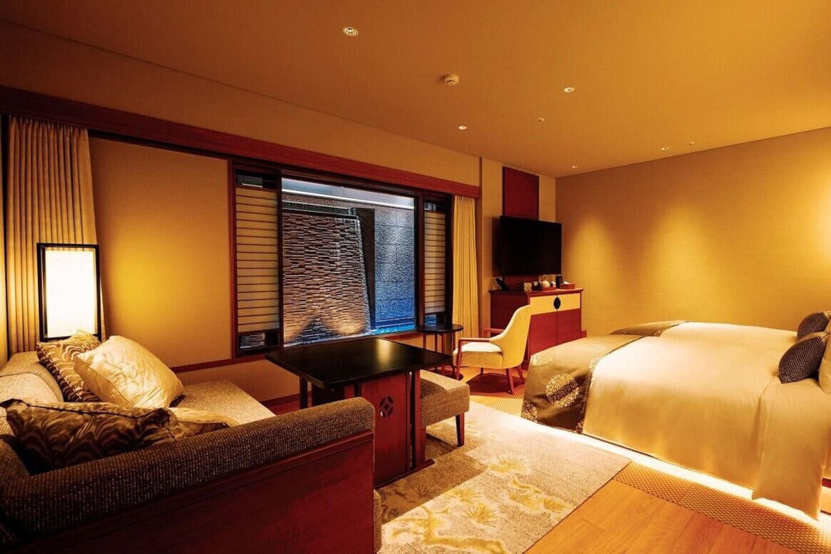 Ein Hotelzimmer mit Bett, Sofa, kleinem Tisch, Fernseher und Fenster mit Jalousien.