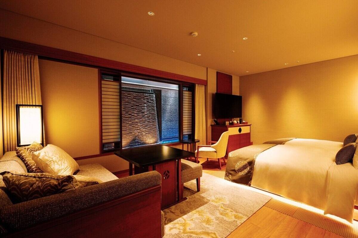 A hotel room with a bed, a sofa, a small table, a TV, and a window with blinds.