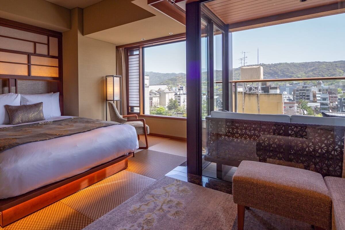 Ein Hotelzimmer mit einem großen Bett, einem Sessel, einem Nachttisch, einer Lampe und Blick auf die Stadt durch große Fenster.