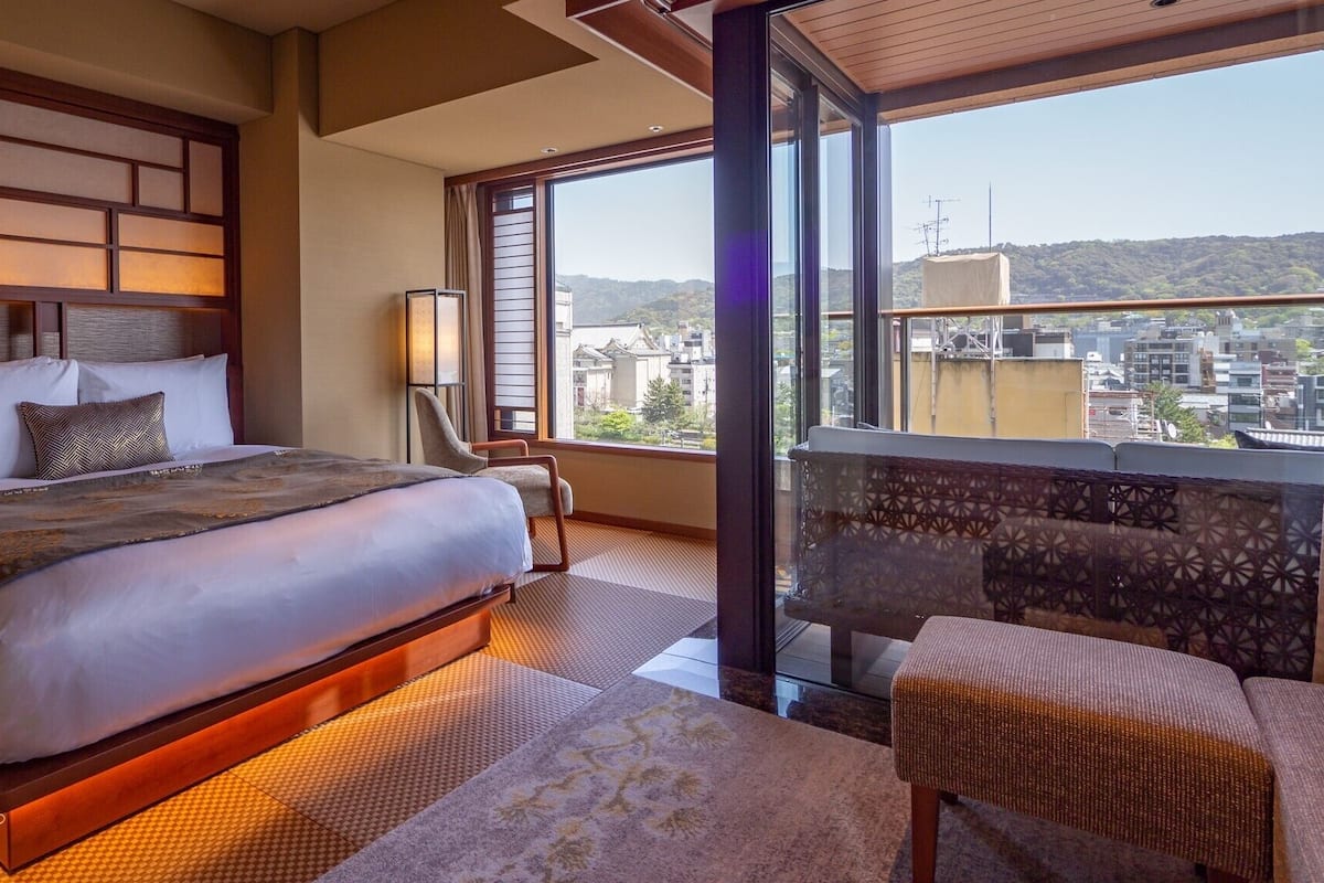 A hotel room with a large bed, a chair, a nightstand, a lamp, and a view of the city through large windows.