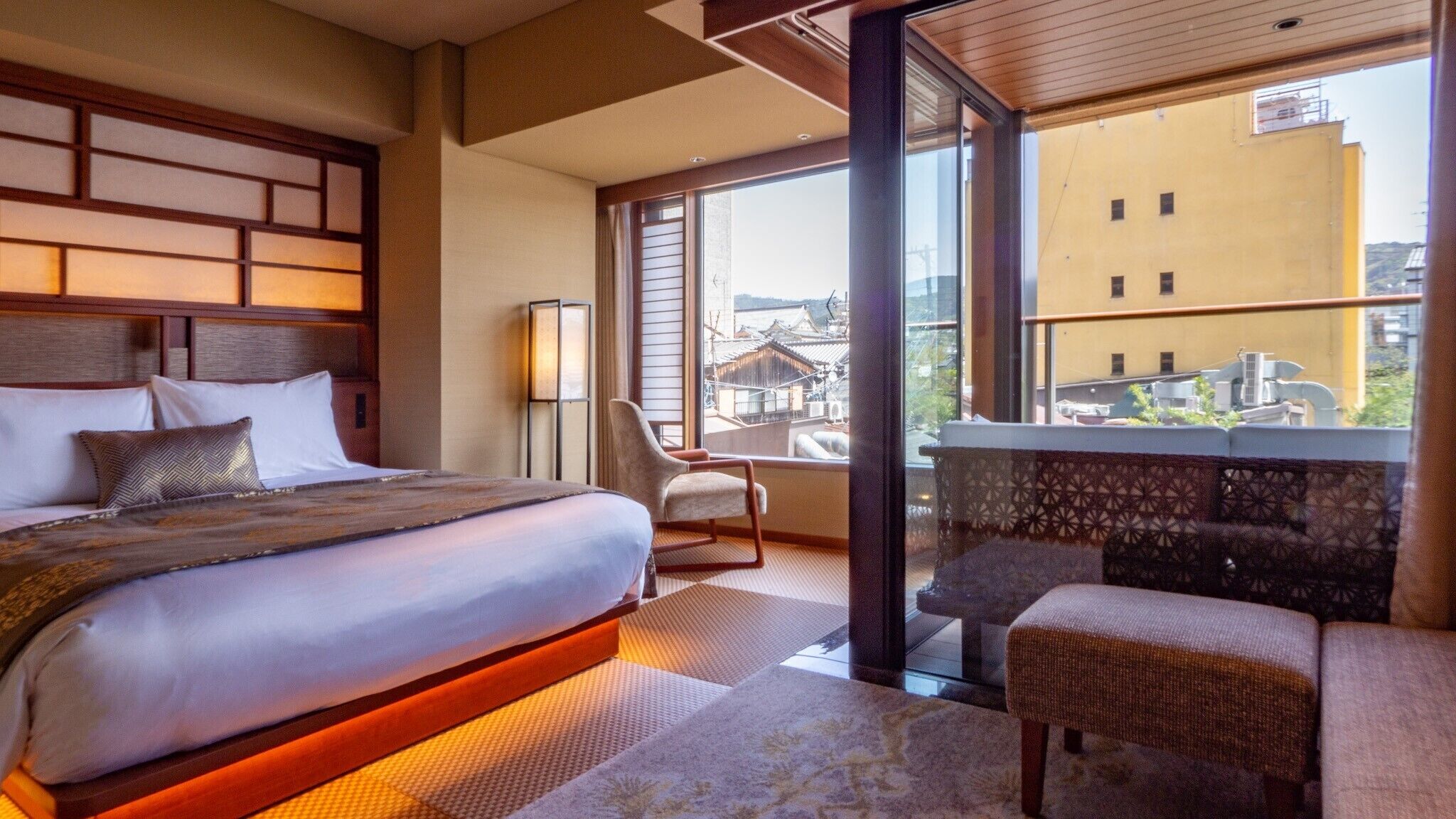 A hotel room with a large bed, a sitting area with a chair and a small table, a balcony with a view of buildings, and a traditional Japanese-style partition.