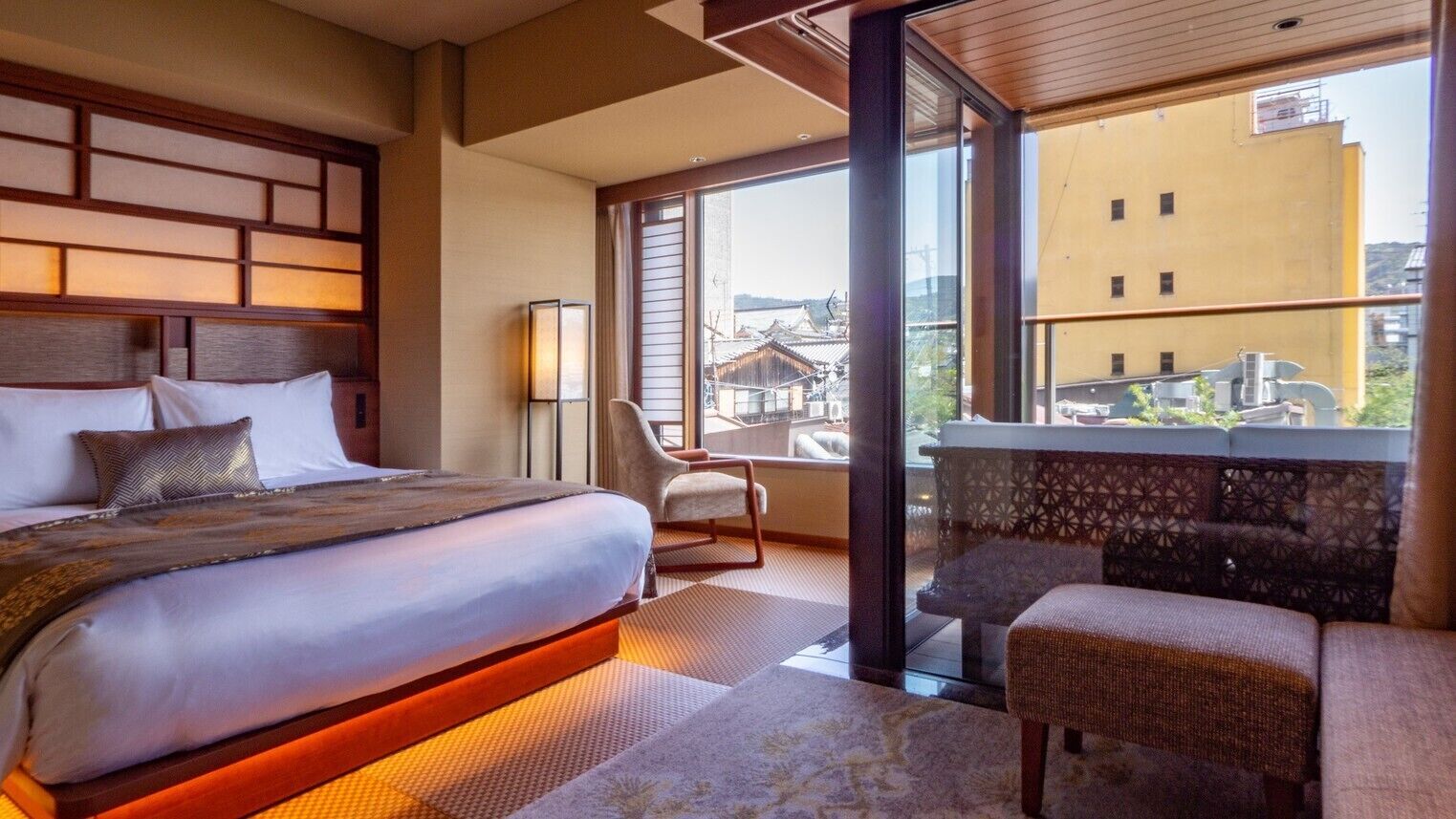 A hotel room with a large bed, a chair, a balcony with a view of buildings, and a patterned wall.