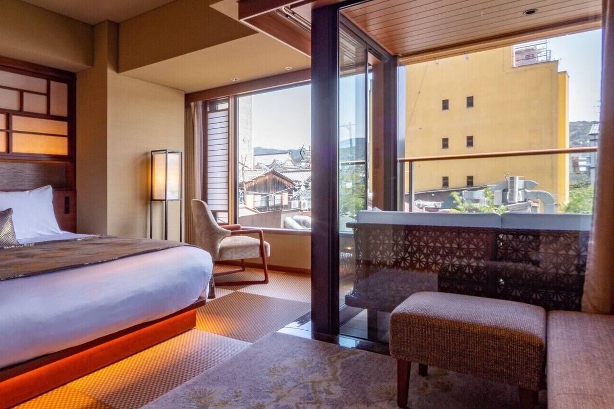 Ein Hotelzimmer mit einem großen Bett, einem Sessel, einem Balkon mit Blick auf Gebäude und einer Wand mit Muster.