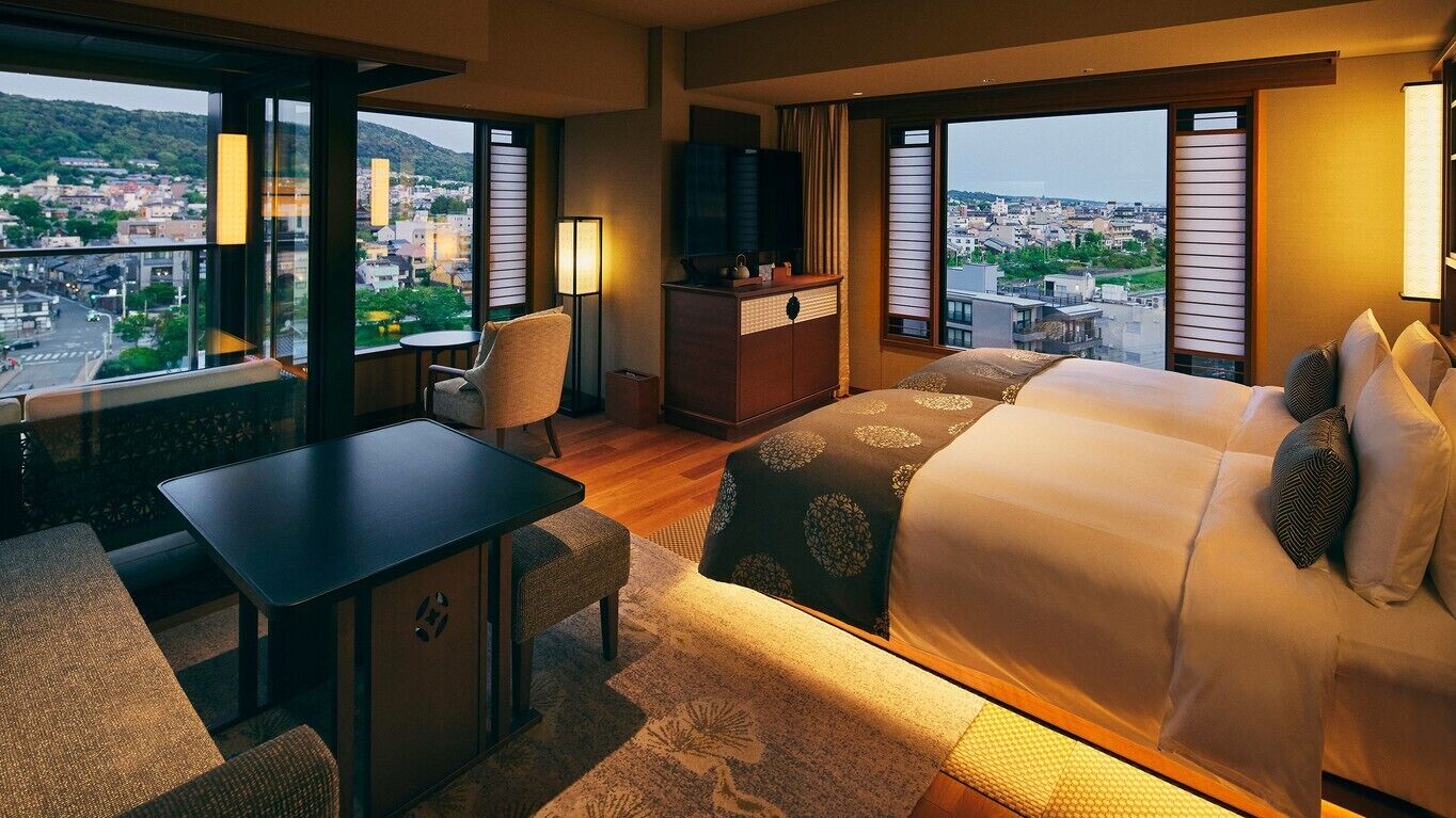 A hotel room with a bed, a desk, a chair, a TV, and large windows with a city view.