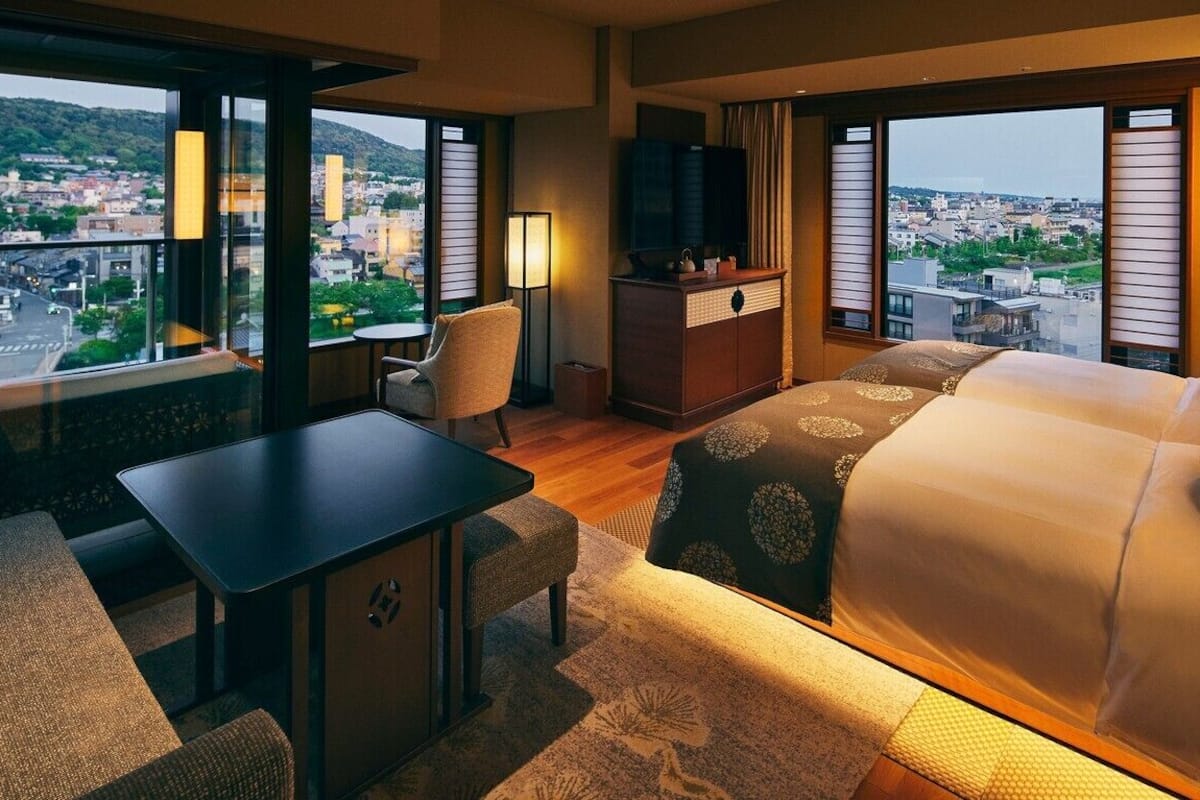 Ein Hotelzimmer mit Bett, Schreibtisch, Stuhl, Fernseher und großen Fenstern mit Blick auf die Stadt.