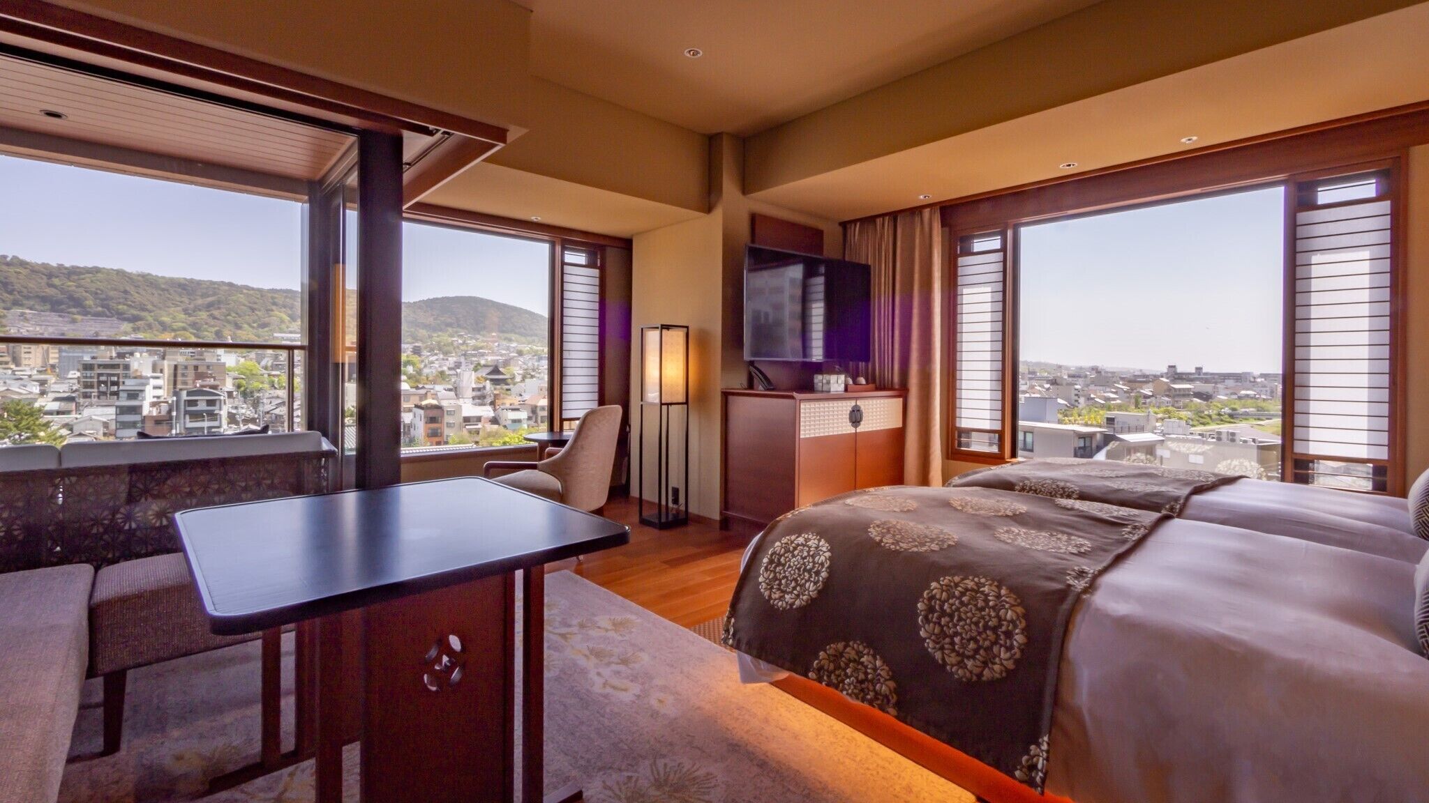 A hotel room with a bed, desk, chair, TV, and large windows with a city view.