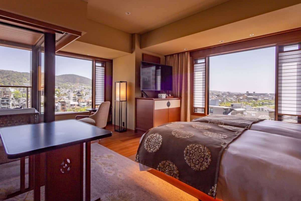 Ein Hotelzimmer mit Bett, Schreibtisch, Stuhl, Fernseher und großen Fenstern mit Blick auf die Stadt.