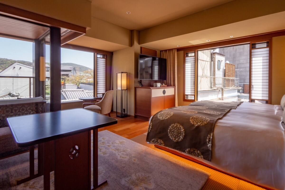 A hotel room with a bed, a desk, a chair, a TV, and a balcony with a view of buildings.
