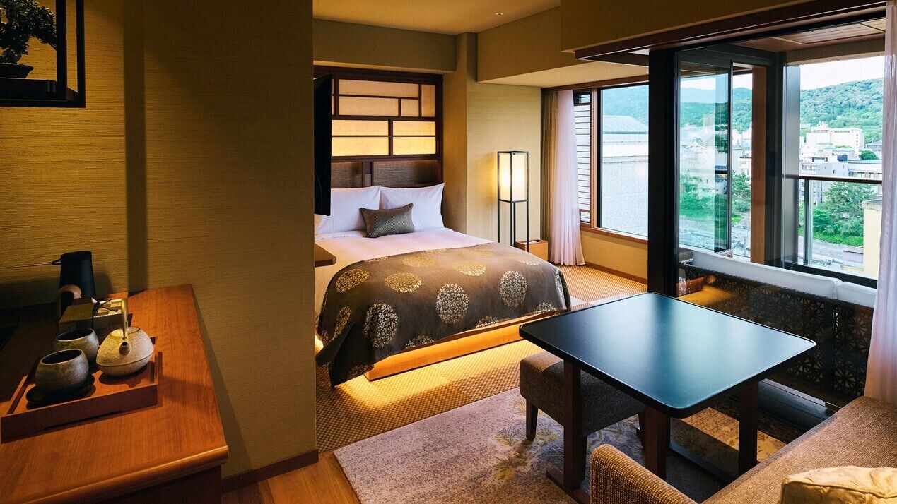 A hotel room with a bed, bedside table, lamp, and a view of the city.