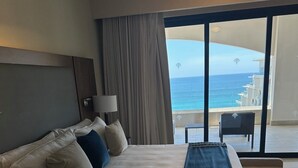 A hotel room with a large bed, a nightstand, a lamp, a view of the ocean through a sliding door, and a chair by the door.