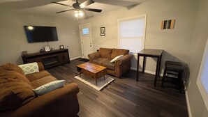 Ein Wohnzimmer mit braunem Sofa, einem Couchtisch, einem Fernseher, einem Deckenventilator und einem Fenster mit Jalousien.