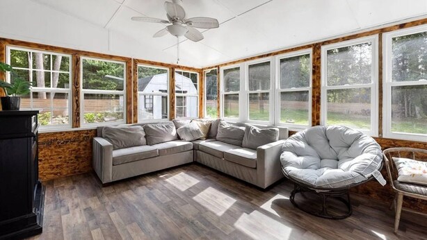 A sunroom with a large window, a ceiling fan, a sectional sofa, a round chair, and a wooden floor. - Virginia poolside Oasis: your Woodland Escape (Chesterfield)