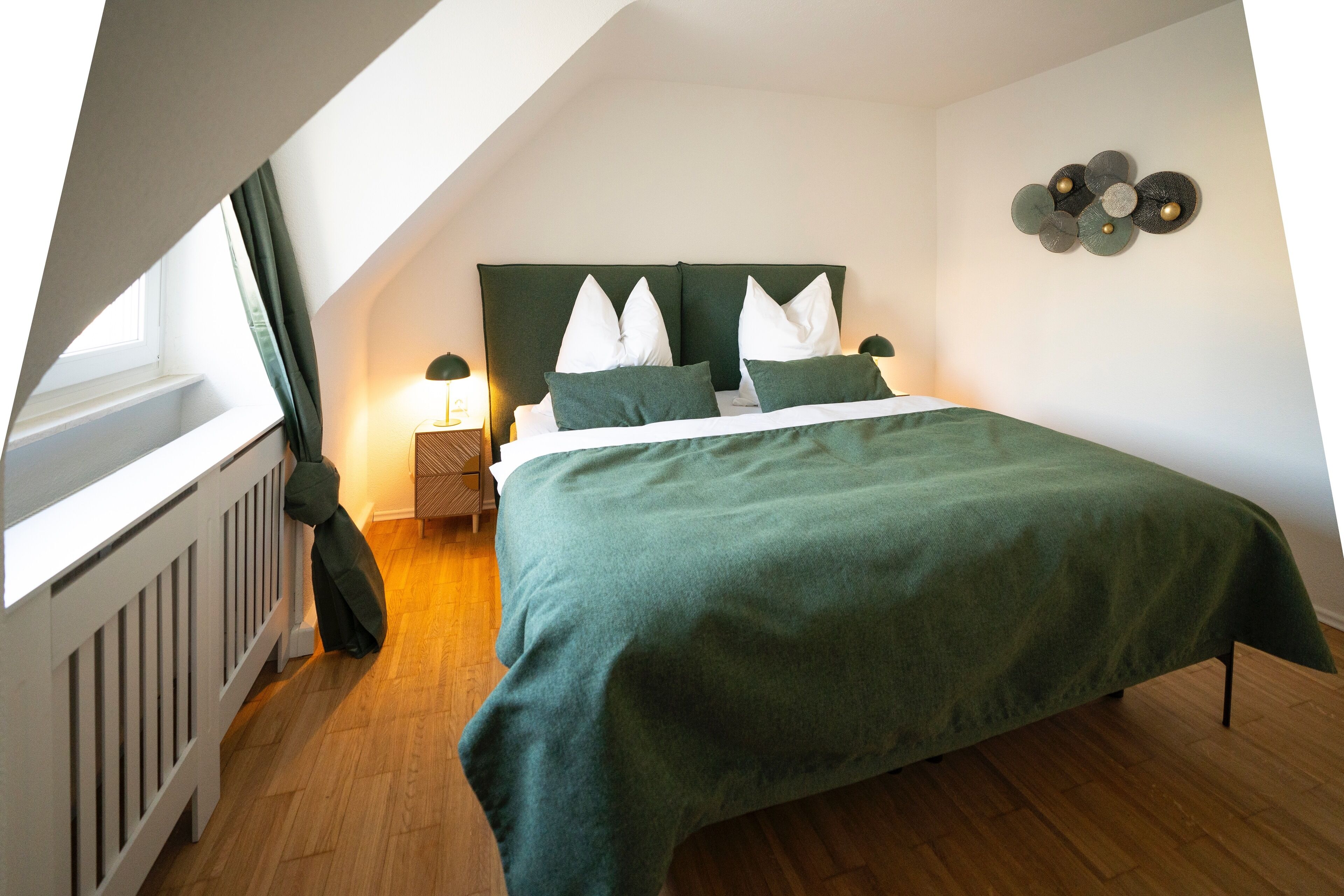 Un dormitorio con techo inclinado, una cama con colcha verde, almohadas blancas, una mesita de noche con lámpara y una decoración en la pared.