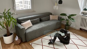 Ein modernes Wohnzimmer mit grauem Sofa, einem Glastisch, einer dekorativen Fischskulptur und einem großen Fenster mit Blick auf Gebäude.
