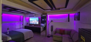 Un dormitorio moderno con cama, sofá, televisión e iluminación púrpura ambiental.