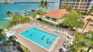A swimming pool surrounded by lounge chairs and palm trees, with a view of the ocean and a building in the background.