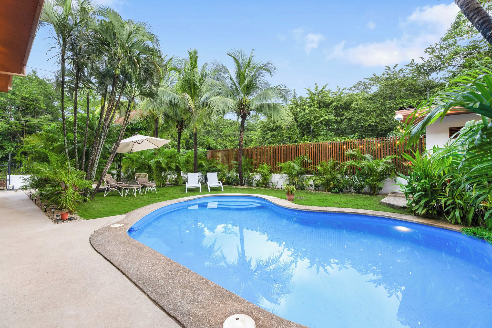 A swimming pool surrounded by lush greenery, with a wooden fence and outdoor seating area.