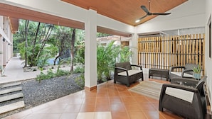 A spacious indoor area with a wooden ceiling, a seating area with wicker furniture, and a view of a tropical garden outside.