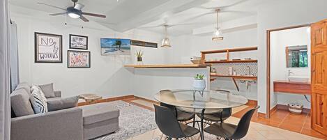 A modern living room with a grey sofa, a glass dining table with black chairs, a ceiling fan, and a kitchen area with a wooden countertop and a sink.