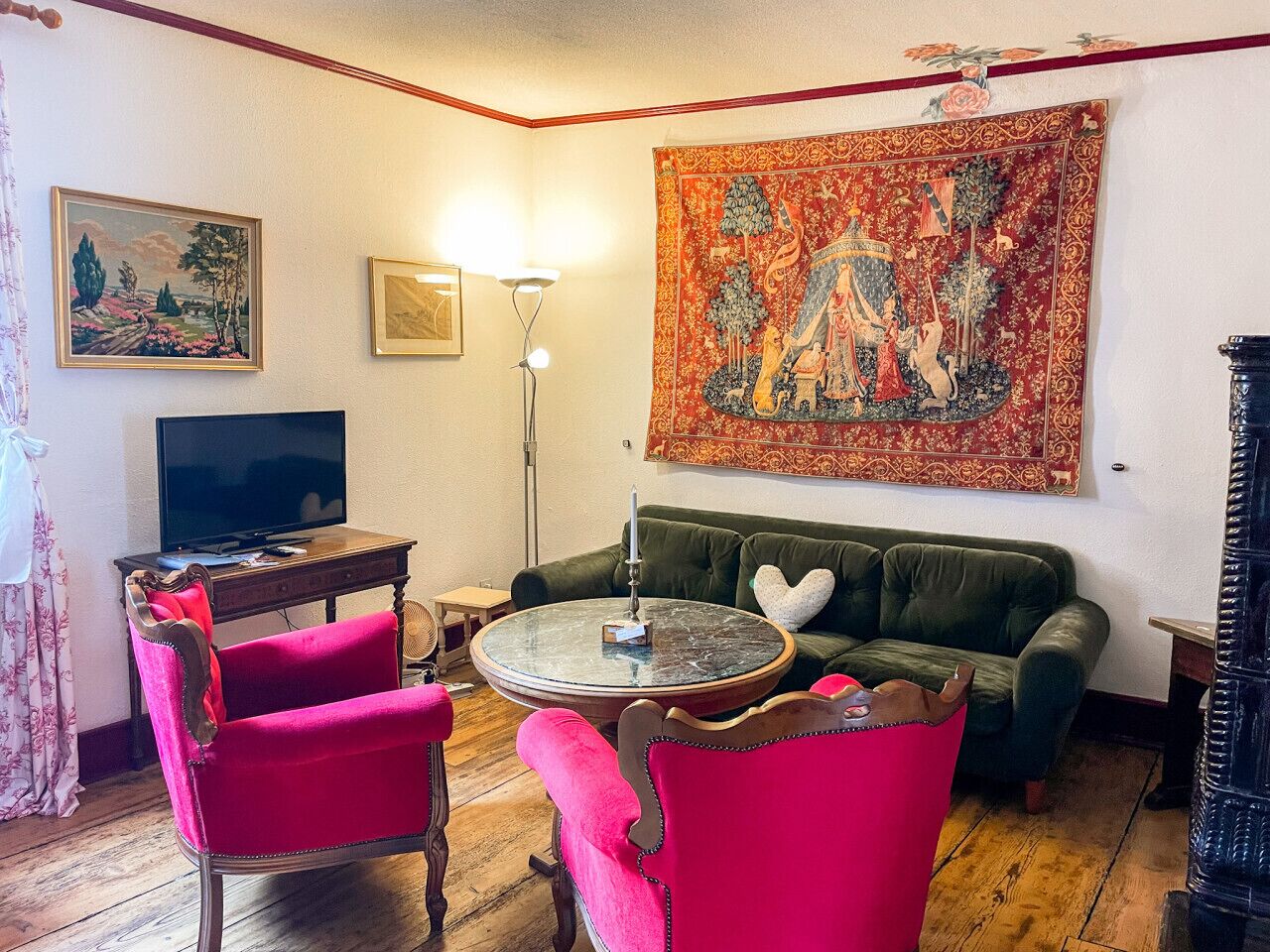 Una sala de estar con un sofá verde, dos sillones rosa, una mesa de centro redonda, un televisor de pantalla plana y un tapiz en la pared.