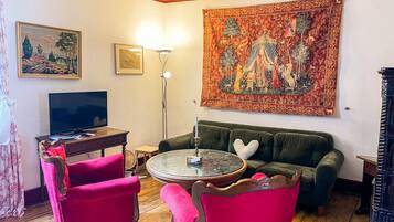 Una sala de estar con un sofá verde, dos sillones rosa, una mesa de centro redonda, un televisor de pantalla plana y un tapiz en la pared.