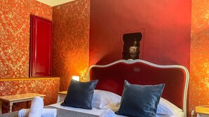 Ein Hotelzimmer mit rotem Kopfteil, zwei Kissen, einem Bett mit grauer Decke und einem kleinen Tischchen mit einem Handtuch.