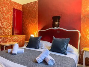Una habitación de hotel con un cabecero rojo, dos almohadas, una cama con una manta gris y una mesita con una toalla.