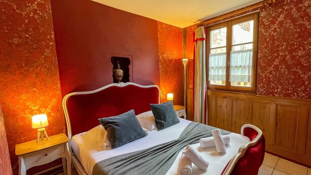 Ein Hotelzimmer mit roter Akzentwand, einem Bett mit weißen Leinentüchern, zwei Kissen, einem Nachttisch mit einer Lampe und einem Fenster mit Vorhängen.